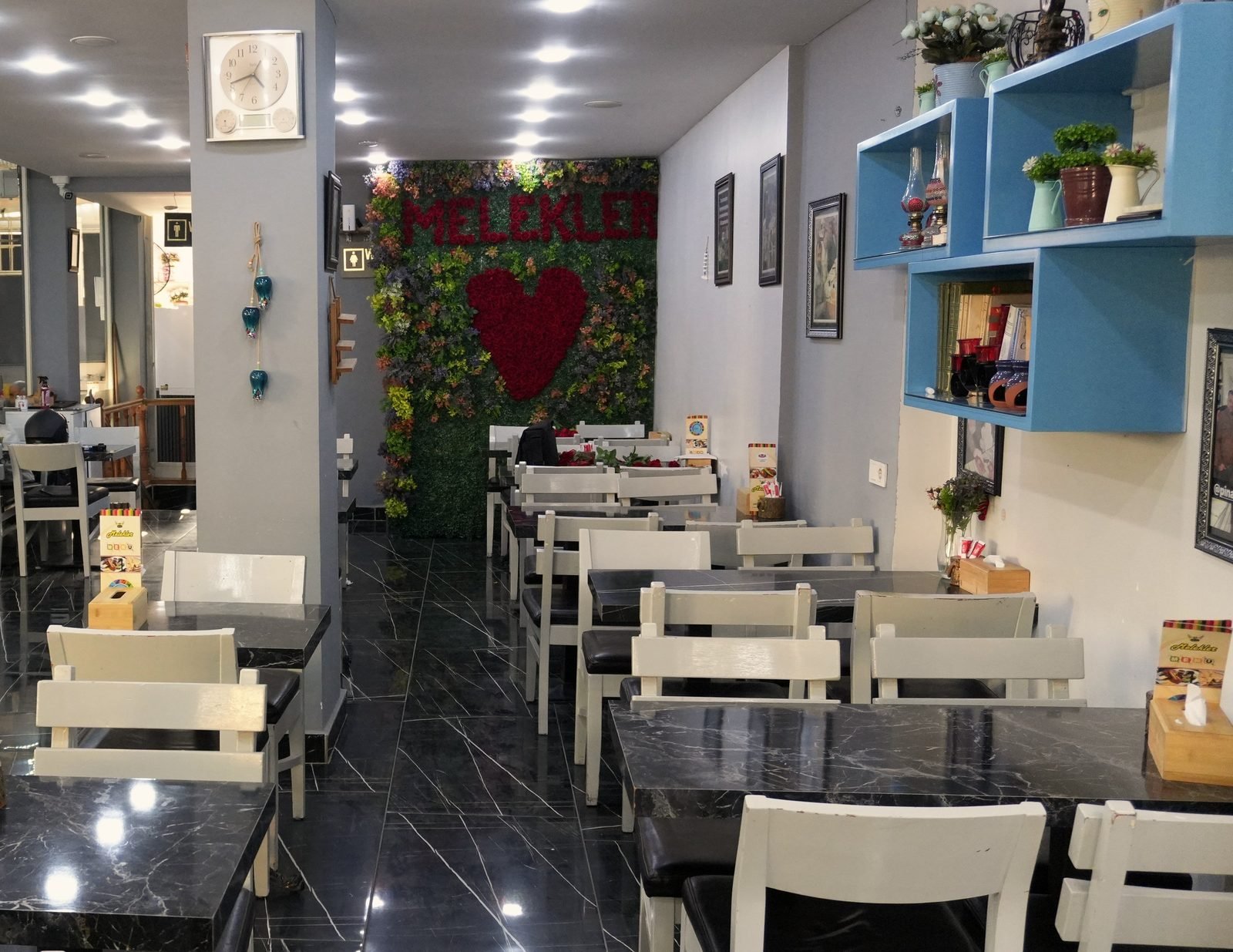 Salon: beyaz sandalyeli mermer masalar, mavi raflar ve arkada kalpli çiçek duvarı