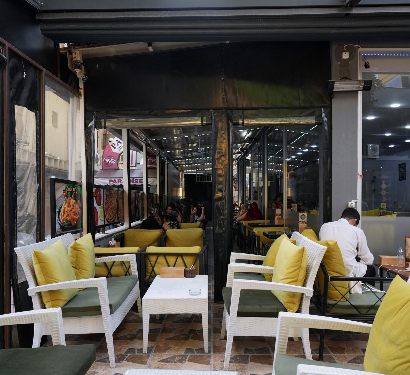 Kapalı teras: sarı minderli hasır koltuklar ve alçak beyaz masalar